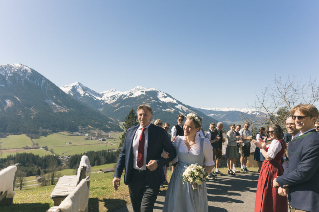 Brautpaar läuft mit Gästen über eine Bergstraße – fröhlicher Auszug nach der Trauung in Kitzbühel