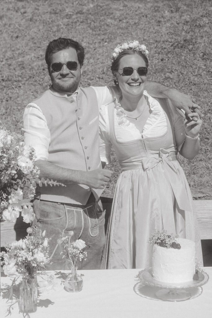 Lachendes Paar mit Sonnenbrille und Dirndl im Sonnenschein – entspannte Hochzeit in Kitzbühel auf der Alm

